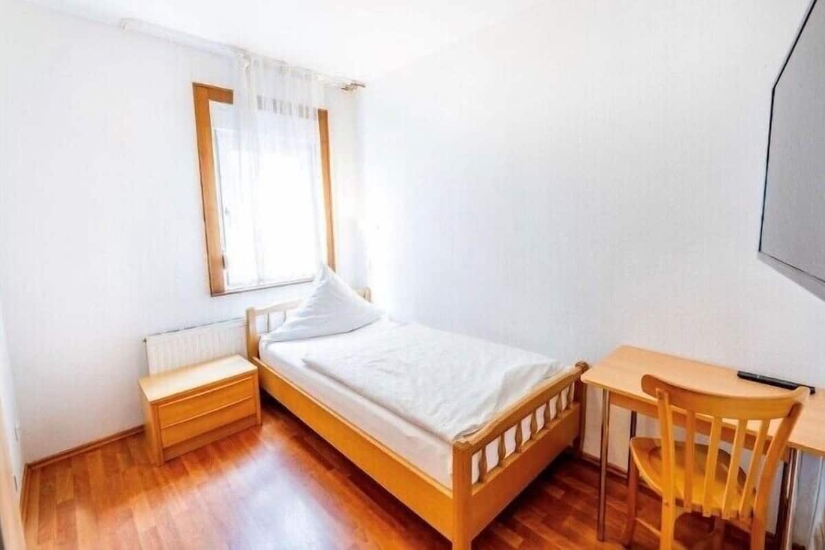 A small, single-bed room with a wooden floor, a nightstand, a desk with a chair, and a window with sheer curtains.