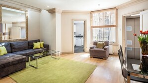 A modern living room with a grey sofa, a glass coffee table, and a green rug. There is a dining table with chairs and a vase of flowers. A large mirror and a window with blinds are also visible. - Chelsea - Draycott Place apartments by Viridian Apartments (London)