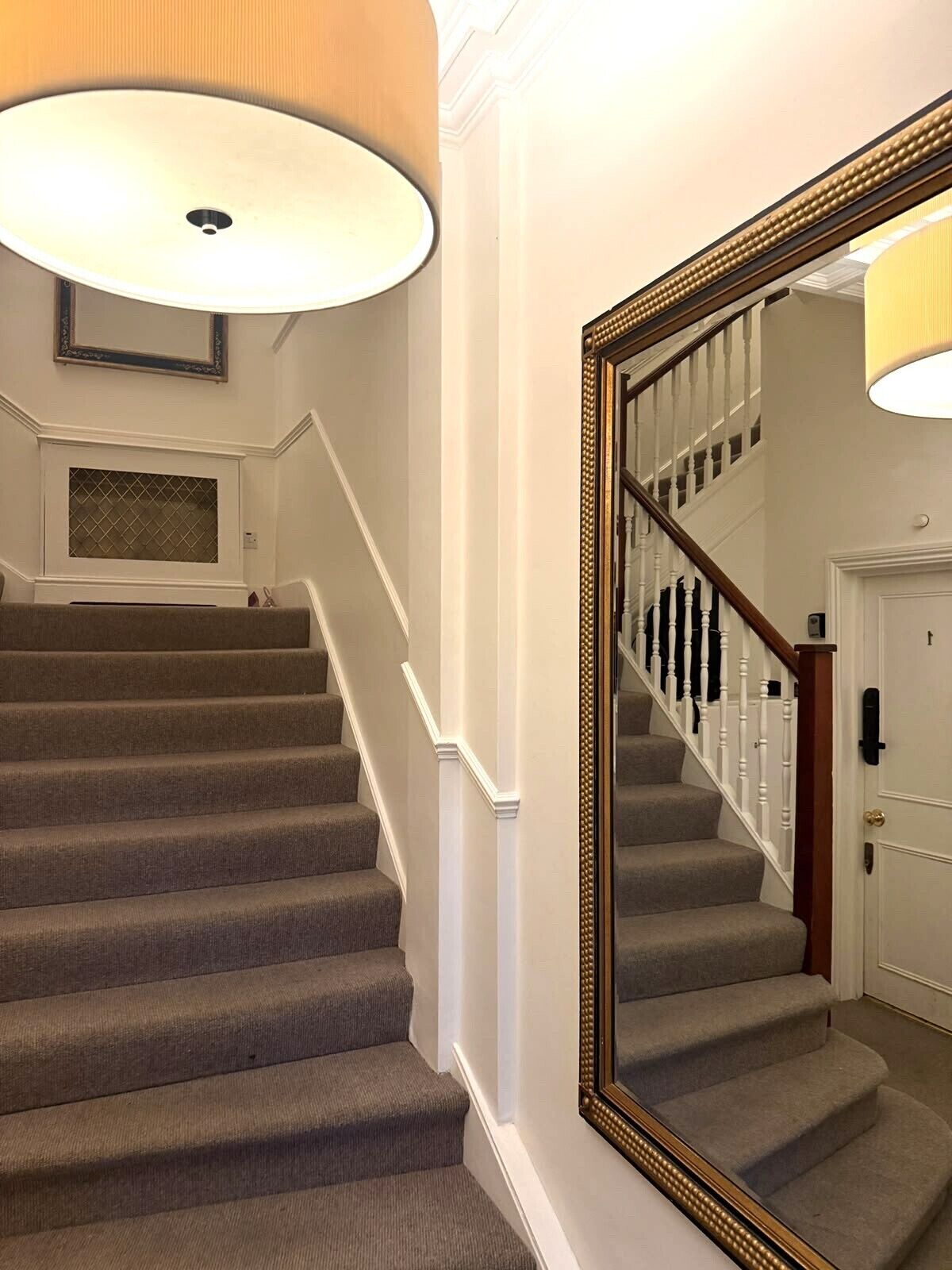 A staircase with carpeted steps, white walls, and a large mirror with a gold frame.