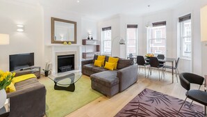 A modern living room with a grey sofa, yellow cushions, a flat-screen TV, a fireplace, and a dining area with a table and chairs. - Chelsea - Draycott Place apartments by Viridian Apartments (London)