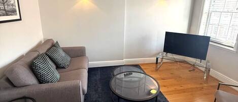 En stue med sofa, sofabord, fjernsyn og et vindue med persienner.