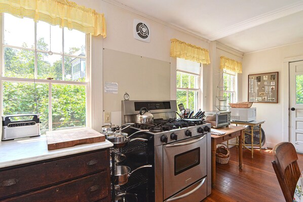 Una cocina con estufa, microondas y diversos utensilios. Hay ventanas con cortinas, un ventilador y un reloj en la pared.