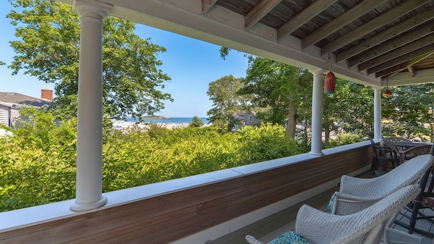 A covered outdoor seating area with white wicker chairs and a wooden bench, overlooking a beach and greenery.