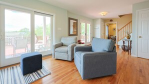 Ein großzügiges Wohnzimmer mit einem großen Fenster, einem grauen Sofa, einem blauen Hocker und einem Holzboden.