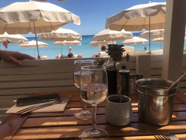 A table with wine glasses, a metal cup, and a small potted plant, set against a backdrop of beach umbrellas and the ocean.