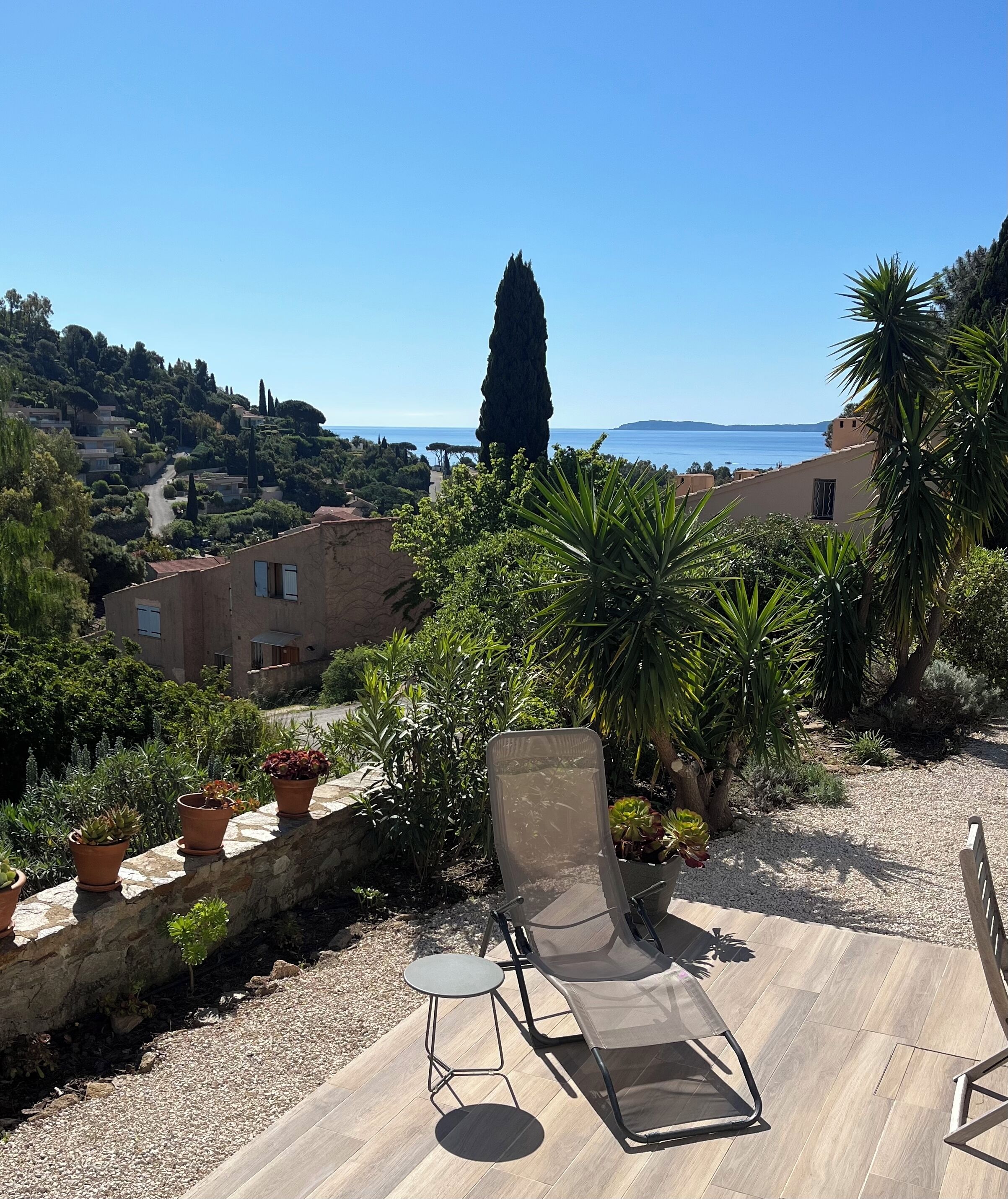 A terrace with a lounge chair, potted plants, and a view of the sea and hillside buildings.