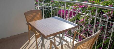 A balcony with a table and two wicker chairs, surrounded by a metal railing and vibrant pink flowers.