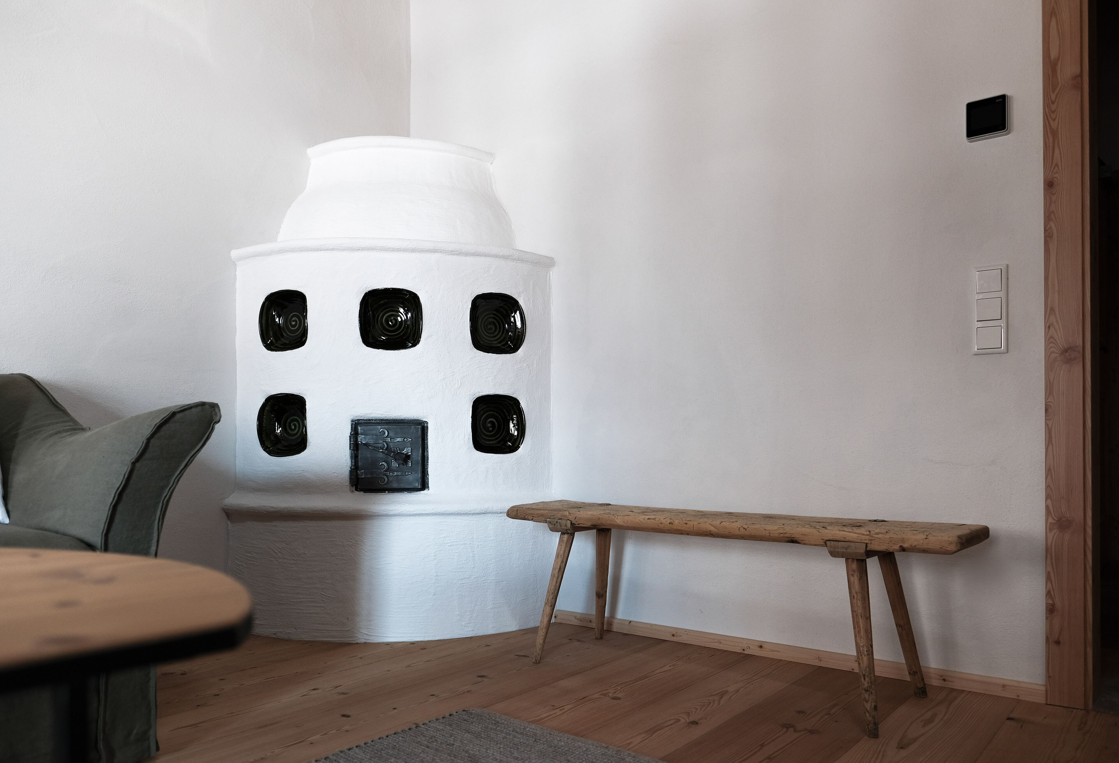 A room with a white, cylindrical, modern fireplace with black circular vents, a wooden bench, a green sofa, and a small table.