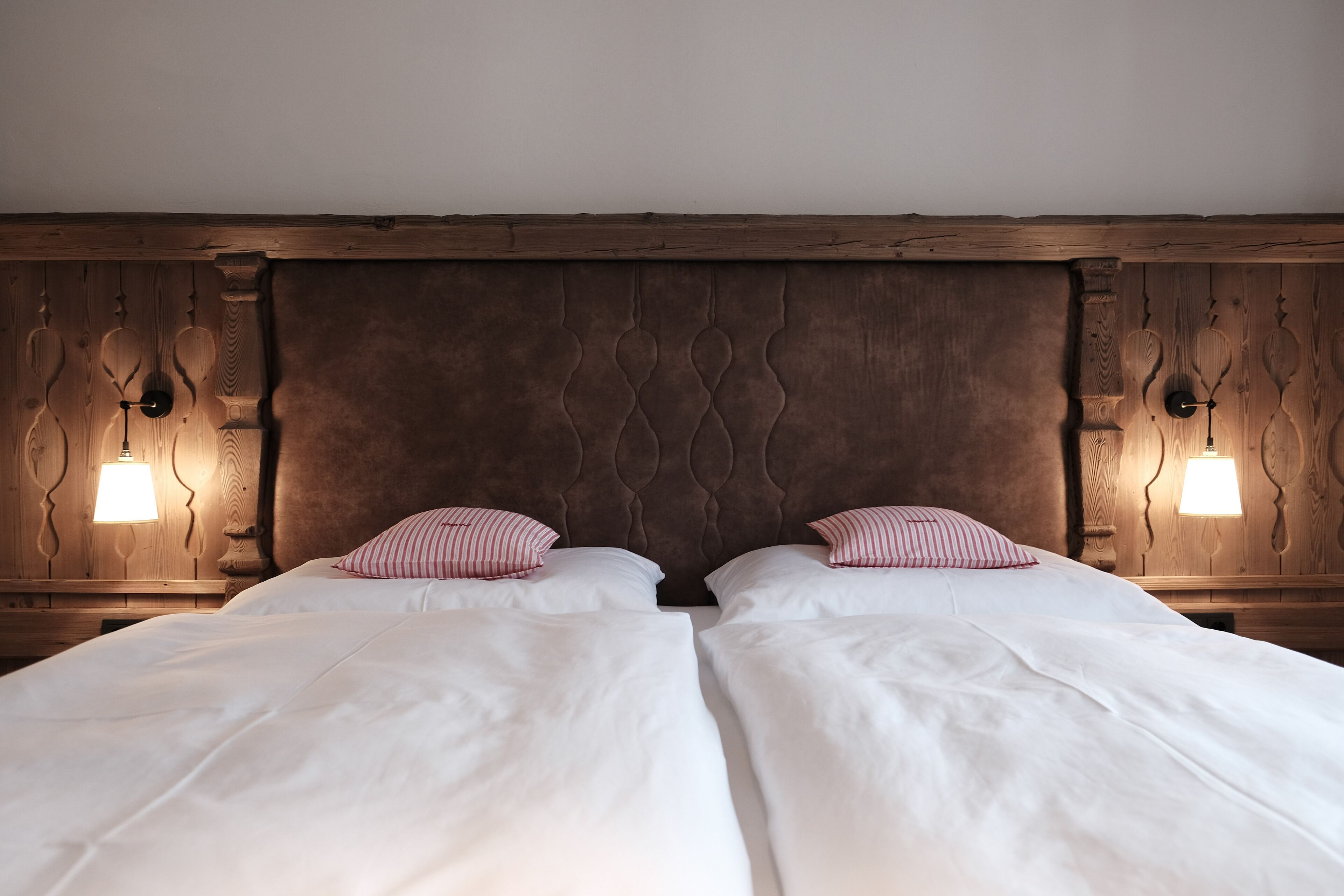 A double bed with white linens and striped pillows, flanked by wooden headboards with wall-mounted lamps.
