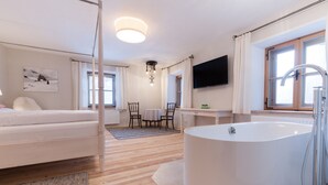 Un dormitorio moderno con una cama grande, una zona de estar con una mesa y sillas, una bañera amplia y un televisor montado en la pared.
