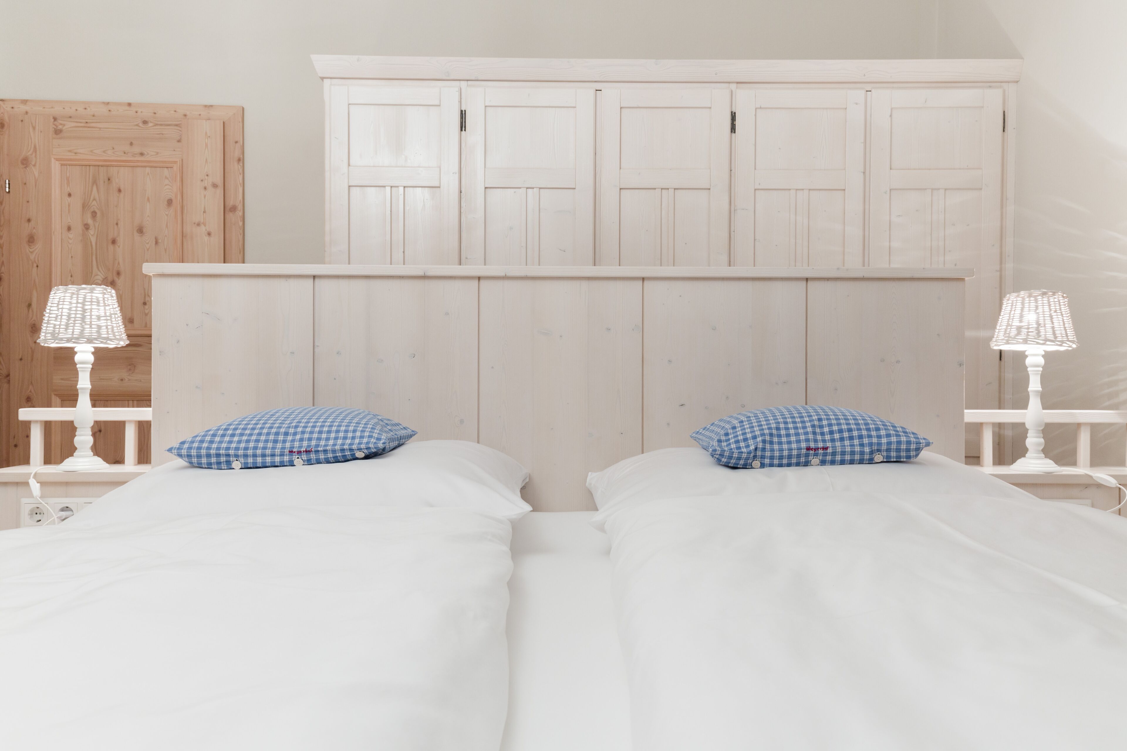 A double bed with white bedding and blue checkered pillows, flanked by white nightstands with lamps, in a room with wooden paneling and a white wardrobe.