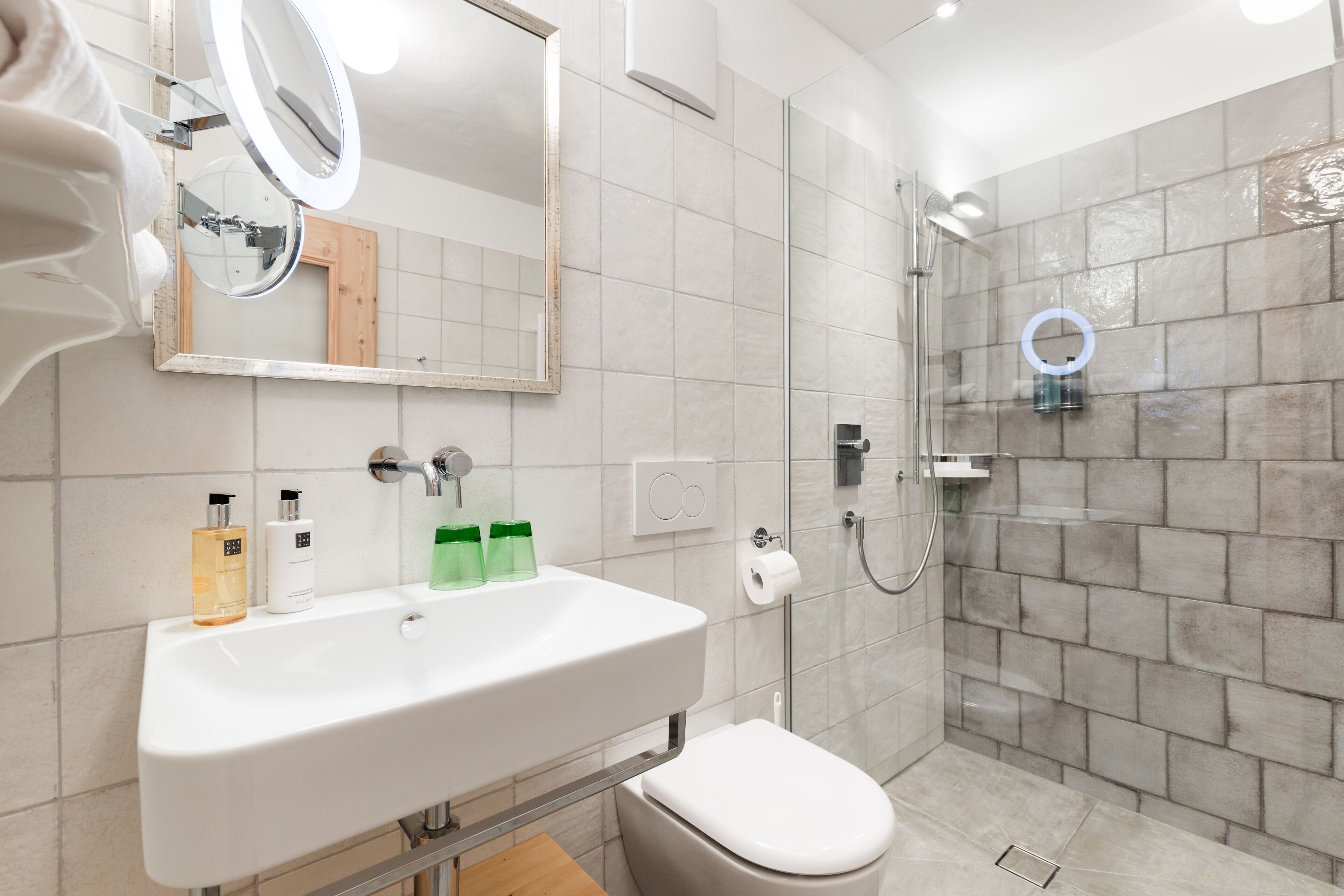 A bathroom with a white sink, mirror, and toilet. It has a shower area with a glass door and a circular mirror on the wall.