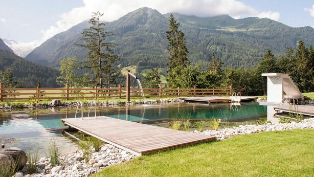 Un paisaje montañoso con una terraza de madera, un estanque pequeño y una construcción de madera.