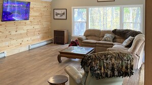 Un salón espacioso con techo de madera, un televisor grande montado en la pared, un sofá color beige, una mesa de centro y una chimenea.