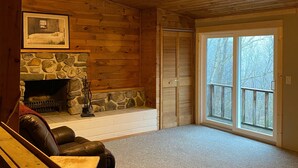 Interior acogedor de una cabaña con chimenea de piedra, ventilador de techo y un gran ventanal corredero que da a una terraza.
