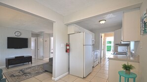 Eine moderne Küche mit weißen Schränken, einem Kühlschrank und einer Spüle. An der Wand hängt eine Uhr und auf einem Hocker steht eine kleine Pflanze.