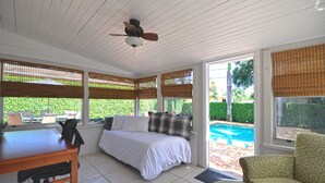 Ein Schlafzimmer mit Bett, Schreibtisch und Stuhl, einem Deckenventilator und großen Fenstern mit Jalousien.