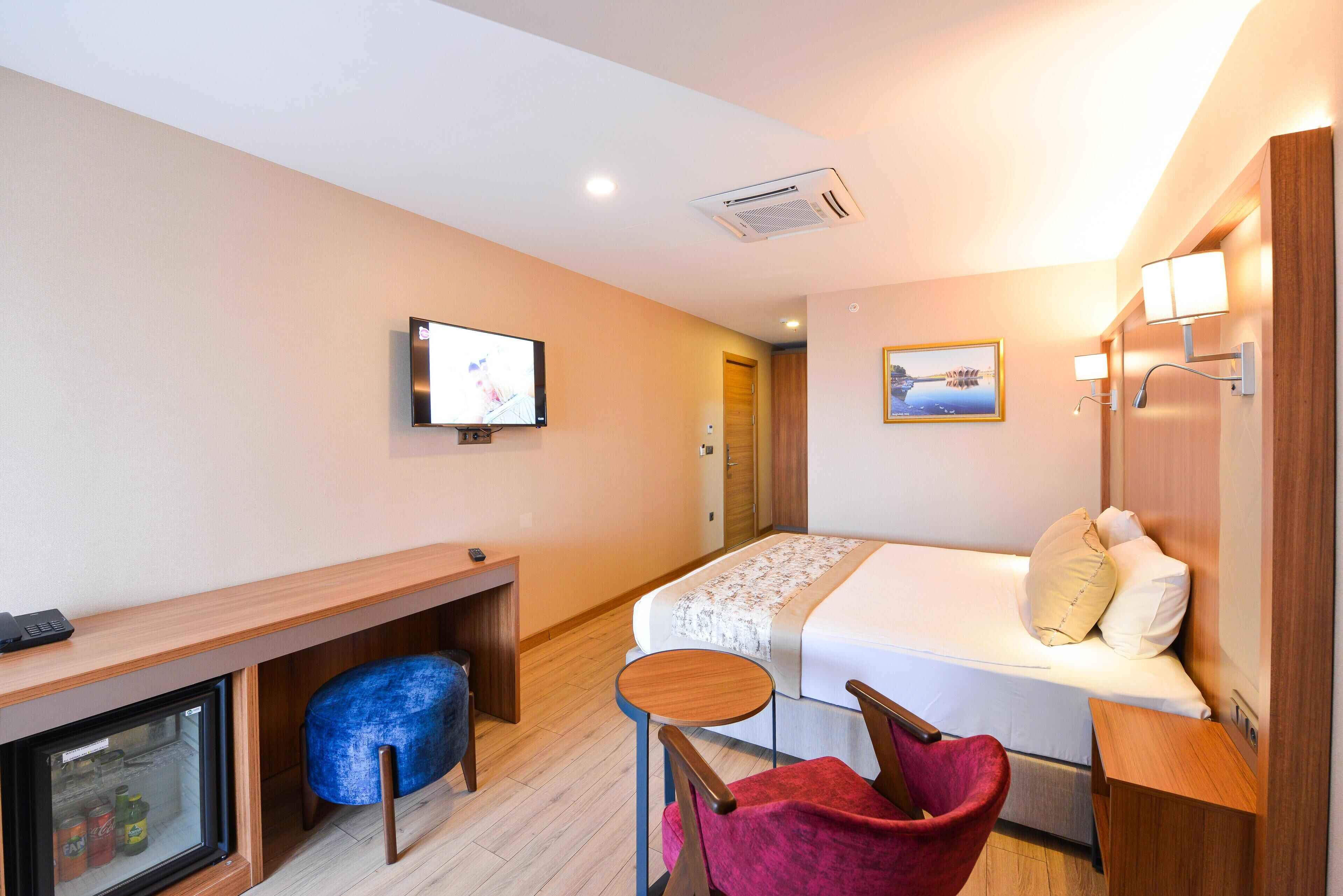 A compact hotel room with a bed, a desk, a chair, a small table, a television, and a refrigerator.