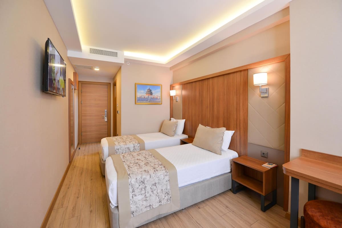 A hotel room with two beds, a wooden headboard, a bedside table, a TV mounted on the wall, and a painting on the wall.
