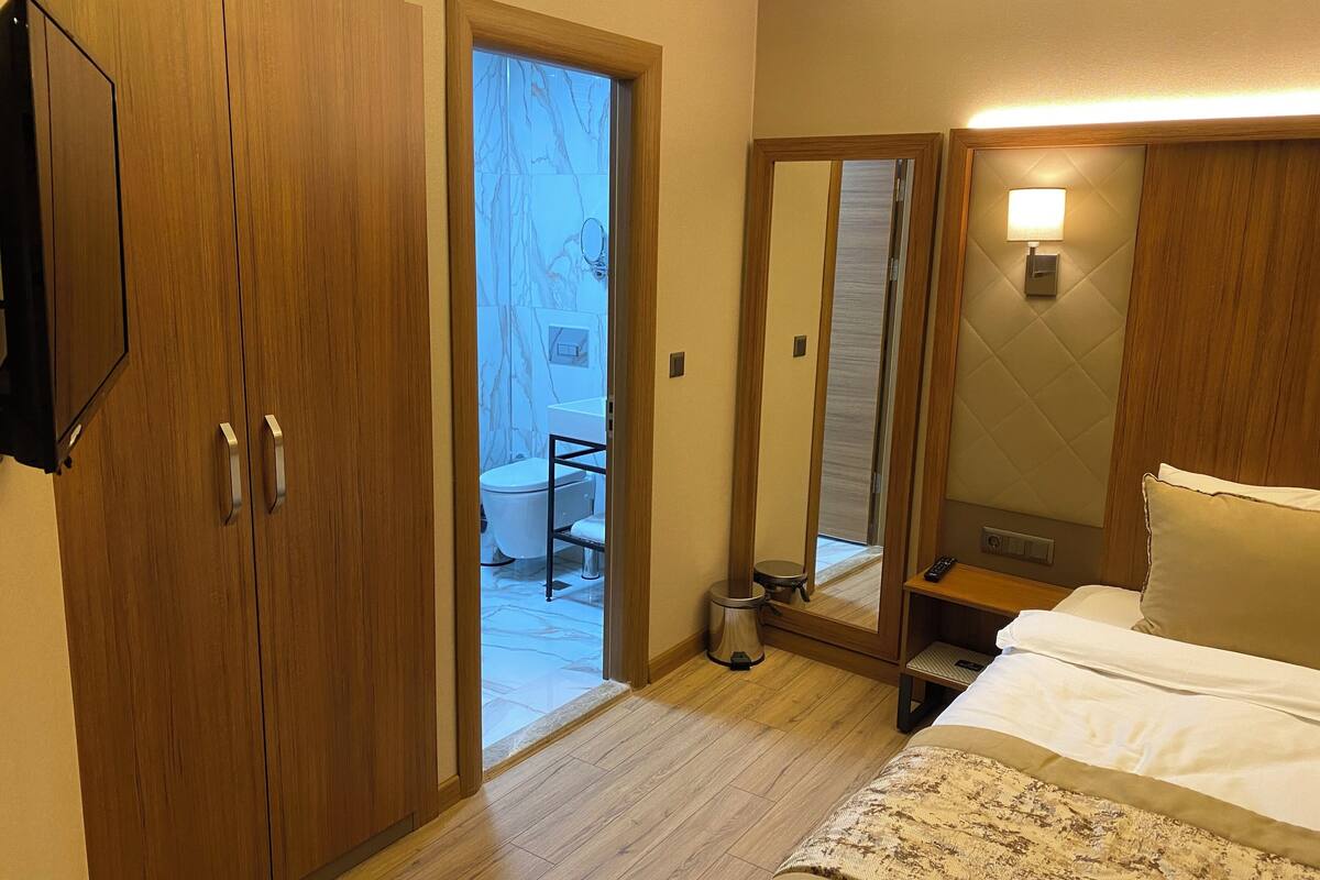 A hotel room with wooden wardrobes, a bed with white linens, a bedside table, a mirror, and a view of the bathroom.