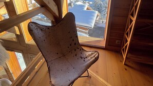 Interno in legno con soffitto alto, un'ampia finestra che mostra un paesaggio montano innevato e una poltrona rustica.