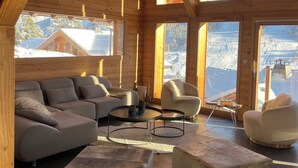 Un ampio interno in legno con un moderno divano, due tavolini rotondi e vista su un paesaggio montano innevato attraverso ampie finestre.