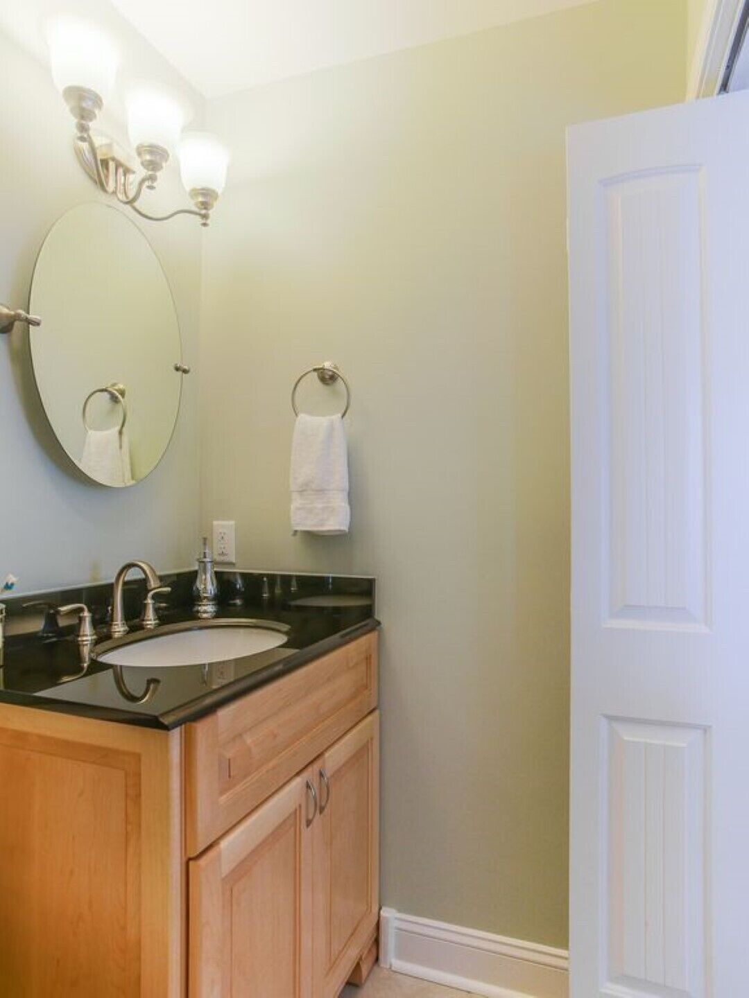 A bathroom with a round mirror, a sink with a black countertop, wooden cabinets, and a towel hanging on a metal ring.