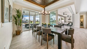 Une salle à manger spacieuse, dotée d’une grande table, de nombreuses chaises et d’un lustre. Des plantes en pot y sont disposées, et de grandes fenêtres offrent une vue sur l’extérieur.