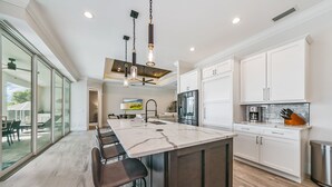 Une cuisine moderne dotée d’un grand îlot central, de meubles blancs et donnant, grâce à des portes vitrées, sur un espace extérieur.