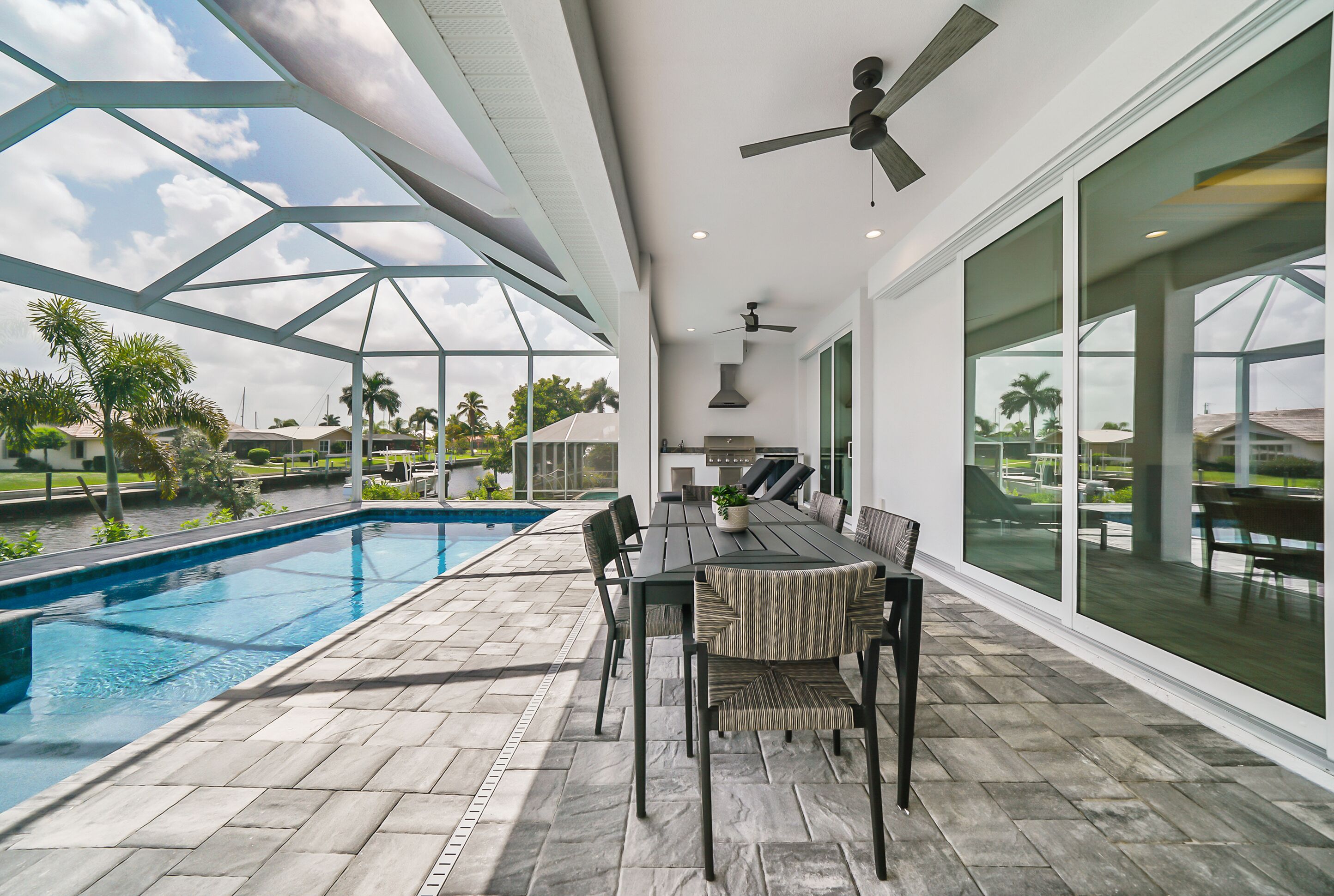 A poolside area with a dining table and chairs, a ceiling fan, and large windows overlooking a swimming pool and greenery.