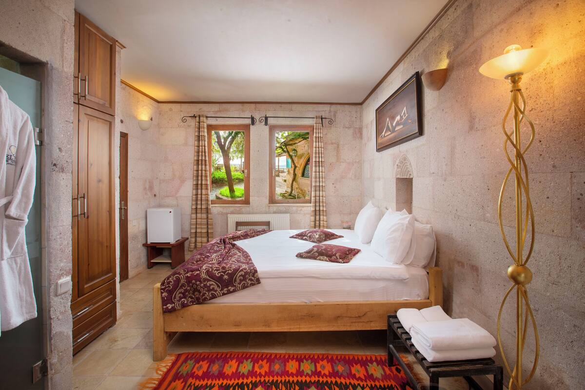 Une chambre avec un lit en bois, une vue sur la verdure à l’extérieur, une lampe fixée au mur et un tableau encadré accroché au mur.