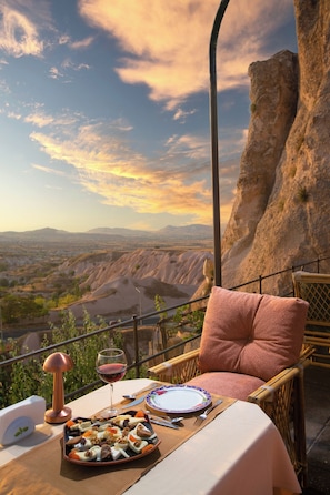 A dining table set with a plate of food, a wine glass, and a chair with a cushion, overlooking a scenic landscape. - Cappadocia Fairy Chimneys Selfie Cave Hotels - Special Class (Nevsehir)
