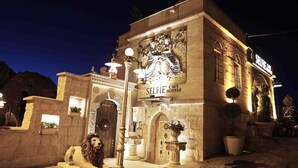 Un bâtiment sur lequel est apposée une enseigne indiquant « SELFIE CAVE HOTELS », illuminé la nuit, avec une architecture en pierre, une statue de lion et des plantes en pot.