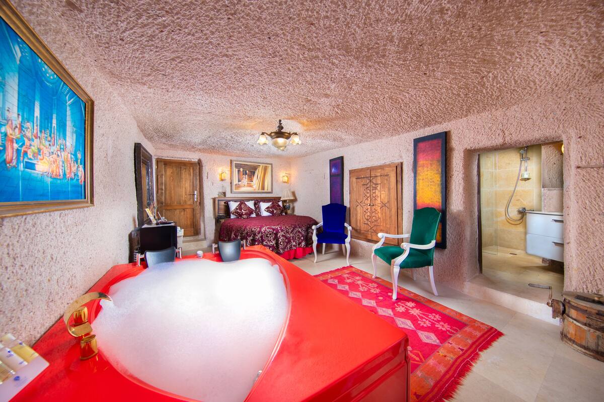 Une pièce avec une grande baignoire rouge, un meuble en bois, un lit recouvert d’une courtepointe rouge et or, et une chaise bleue.