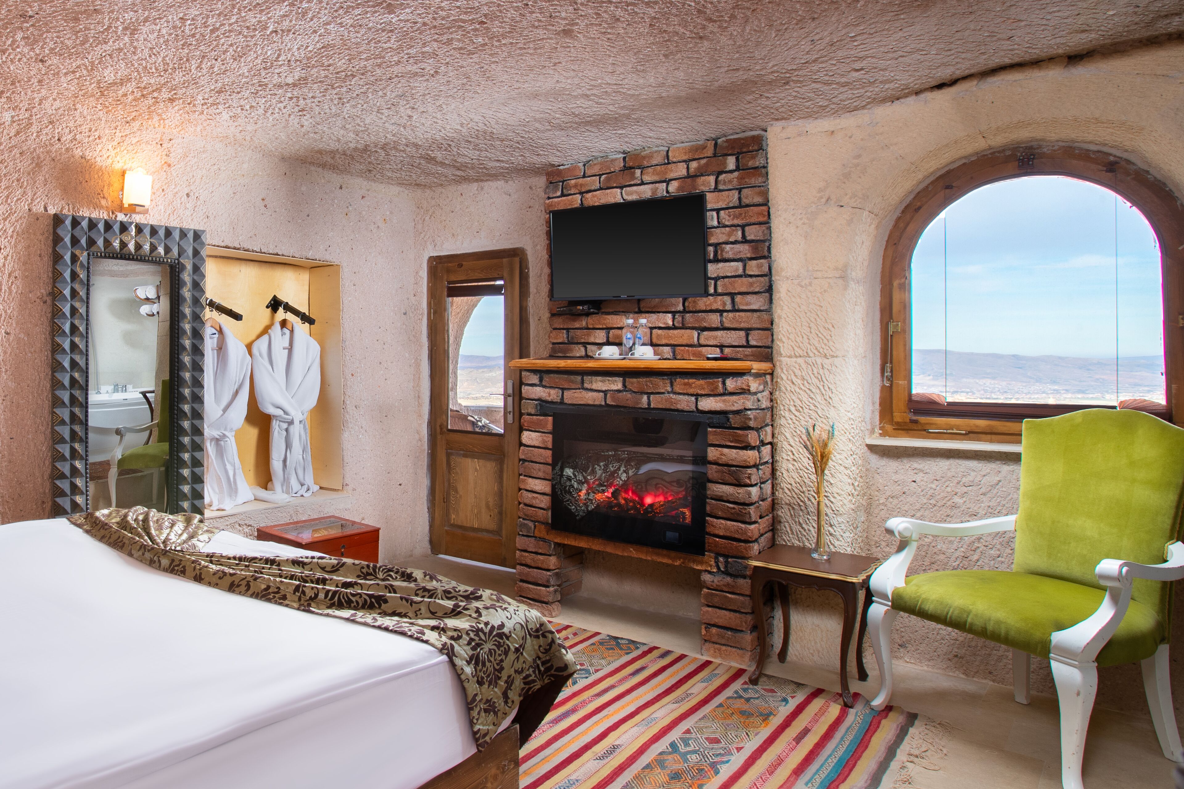 A room with a brick fireplace, a bed, a green chair, and a large arched window offering a view.