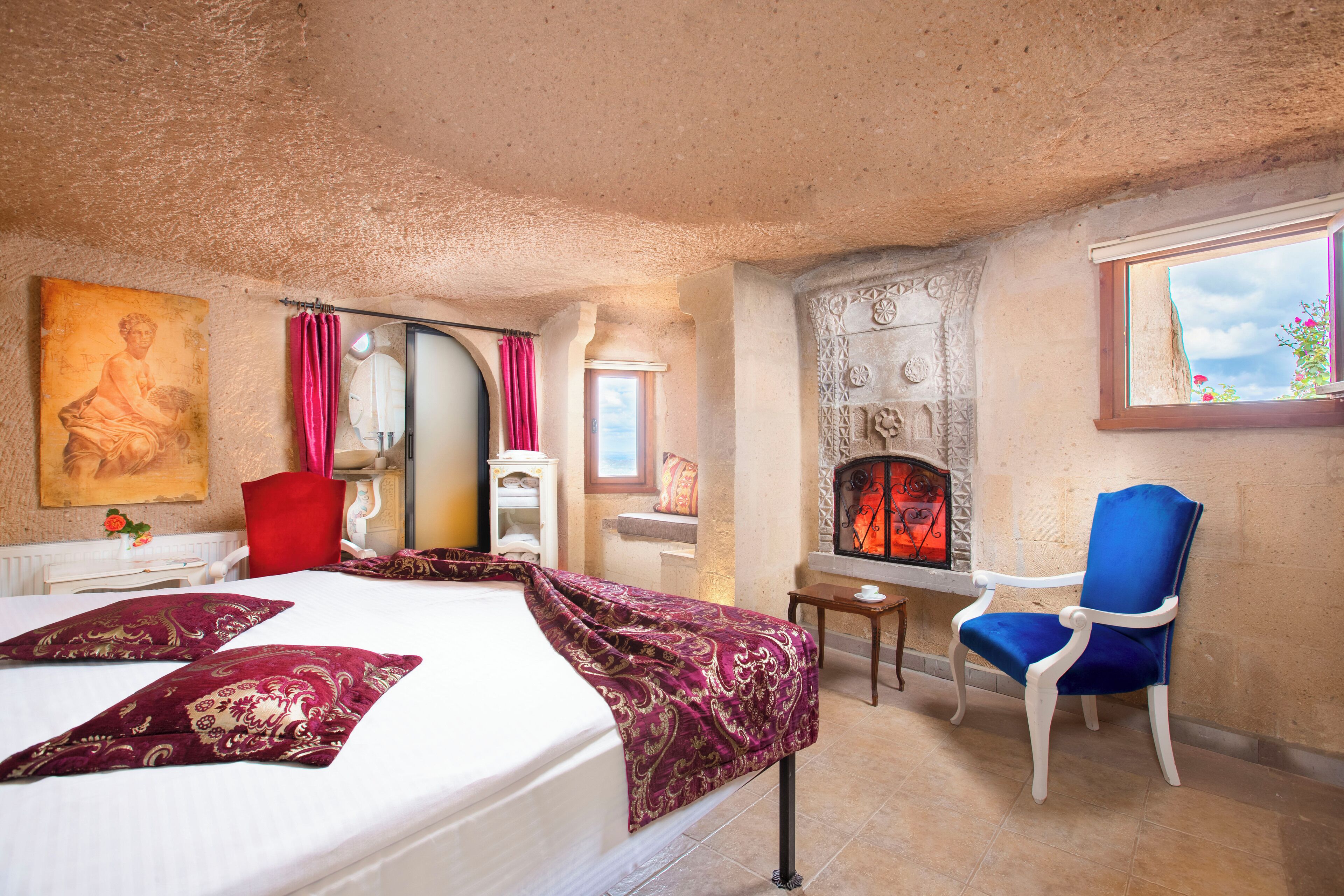 A bedroom with a bed, a blue chair, a fireplace, and a painting on the wall.