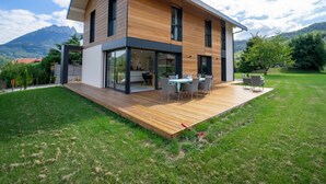 A modern two-story house with a wooden deck, large windows, and a view of mountains.