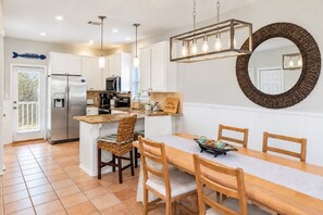 A kitchen with white cabinets, a stainless steel refrigerator, a microwave, and a sink. There is a dining table with chairs and a large mirror on the wall.