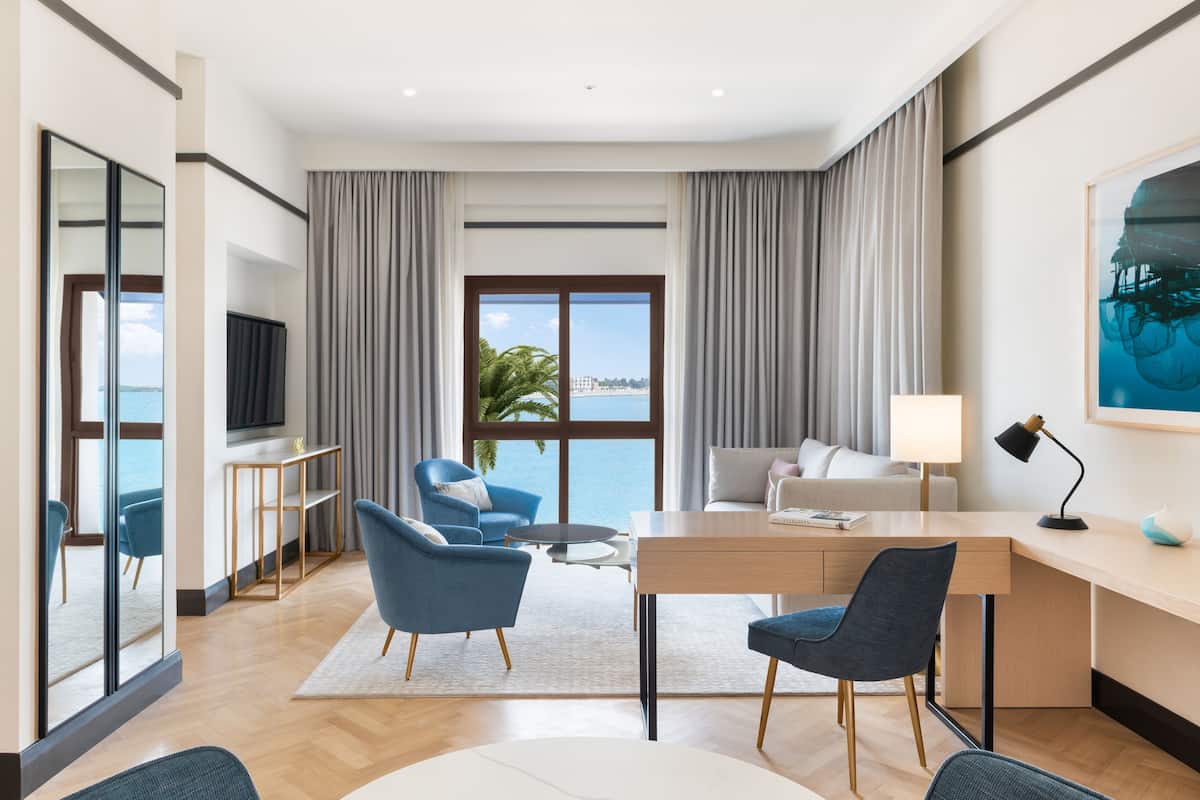 Ein modernes Hotelzimmer mit großem Fenster, Schreibtisch, zwei blauen Sesseln, einer Couch und einem Fernseher an der Wand.