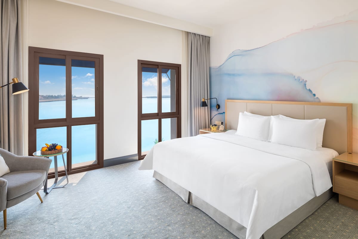 Ein Hotelzimmer mit einem großen Bett, einem Sessel, einem kleinen Tisch mit Früchten und Meerblick durch große Fenster.