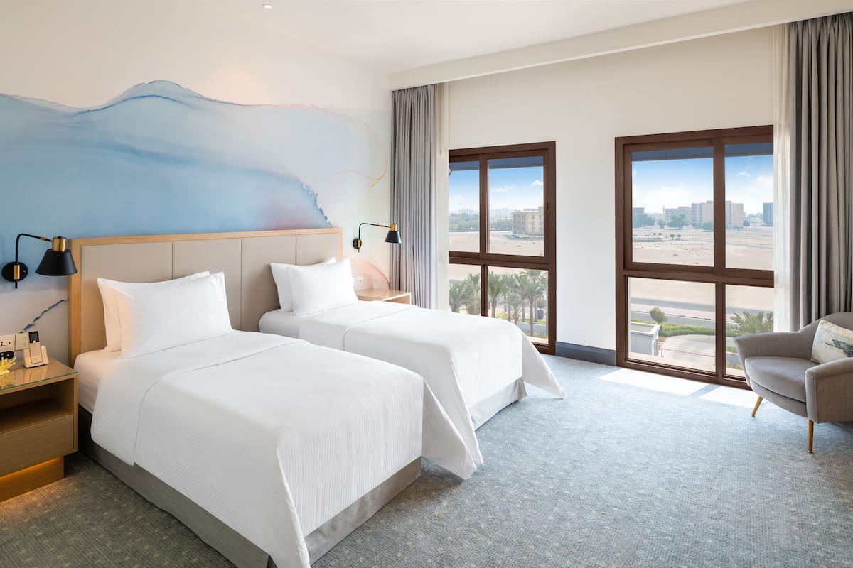 Ein Hotelzimmer mit zwei Betten, einem Schreibtisch, einem Stuhl und großen Fenstern mit Blick auf die Stadt.