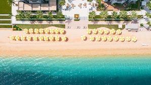 Vista aérea de una playa con hileras de sombrillas y sillas de playa, un área de piscina con terraza, y un edificio de diseño moderno.