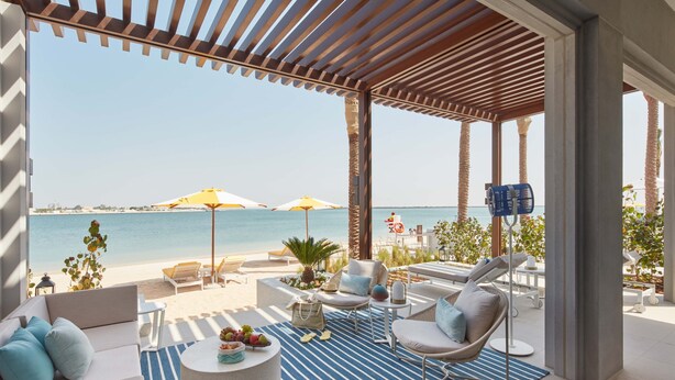 Un área de descanso junto a la playa con vista al mar, que incluye un sofá, sillas, una mesa redonda y sombrillas.