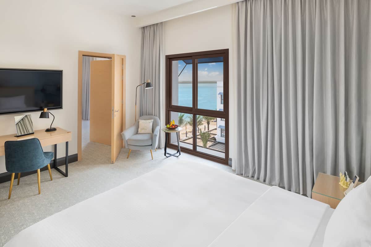 Ein Hotelzimmer mit Bett, Schreibtisch inklusive Computer, Stuhl, Fernseher, Fenster mit Meerblick und einem kleinen Tisch mit Früchten.