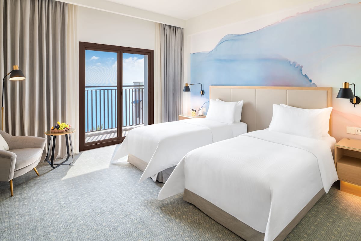 Ein Hotelzimmer mit zwei Betten, einem Sessel, einem kleinen Tisch mit Obst und einem Balkon mit Blick.