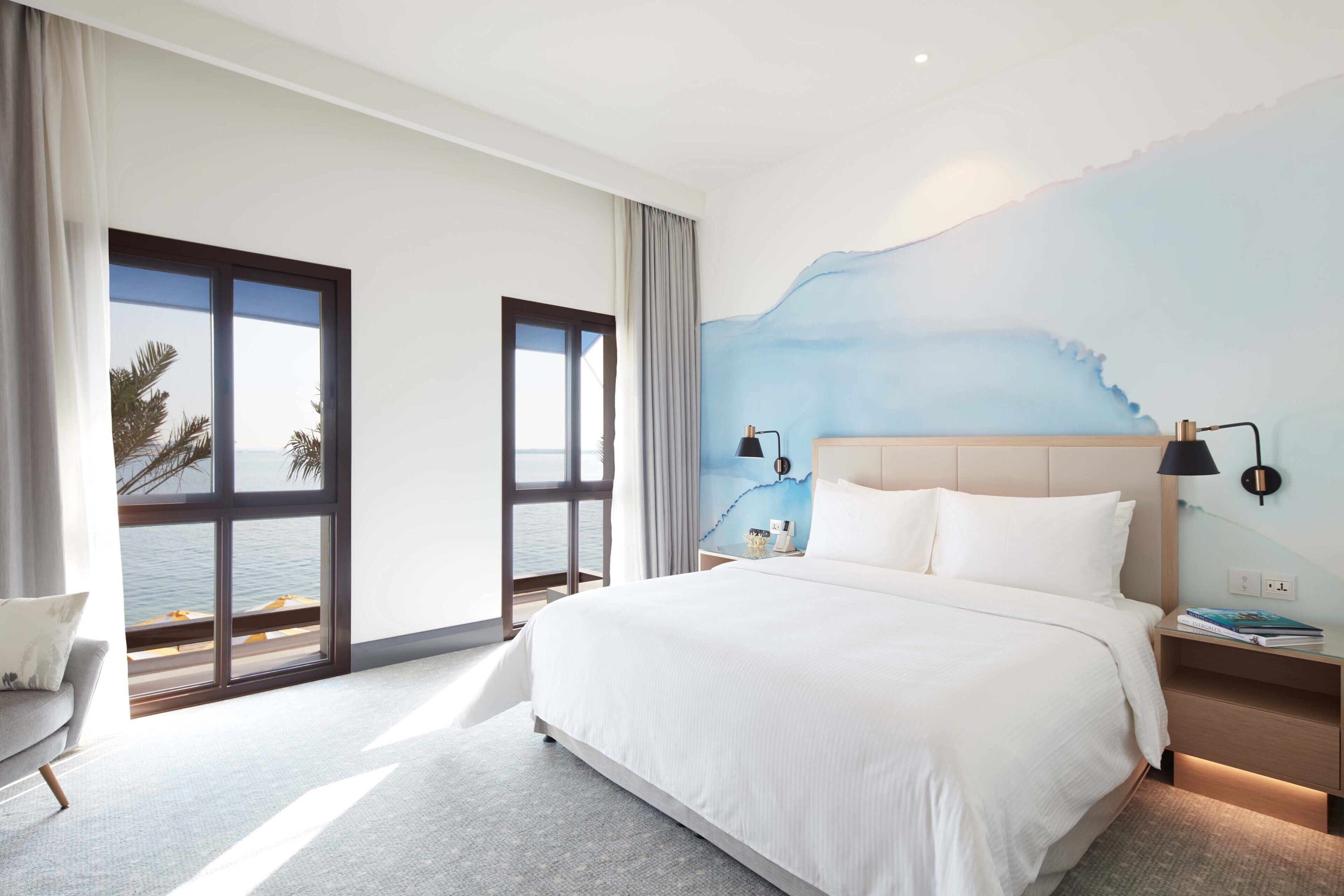 A modern hotel room with a large bed, a view of the sea through floor-to-ceiling windows, a wall mural, and a small seating area.