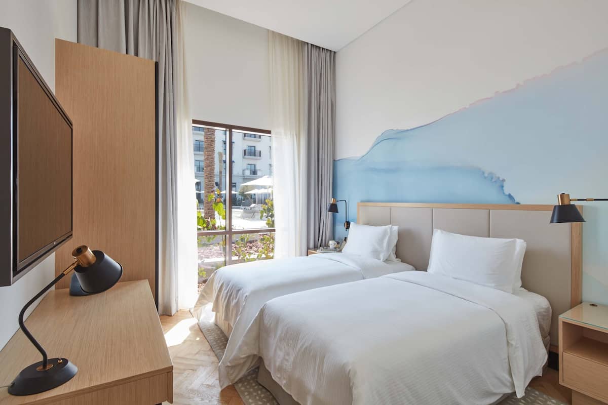 Ein Hotelzimmer mit zwei Betten, einem Schreibtisch, einem Fernseher und einem großen Fenster mit Blick nach draußen.