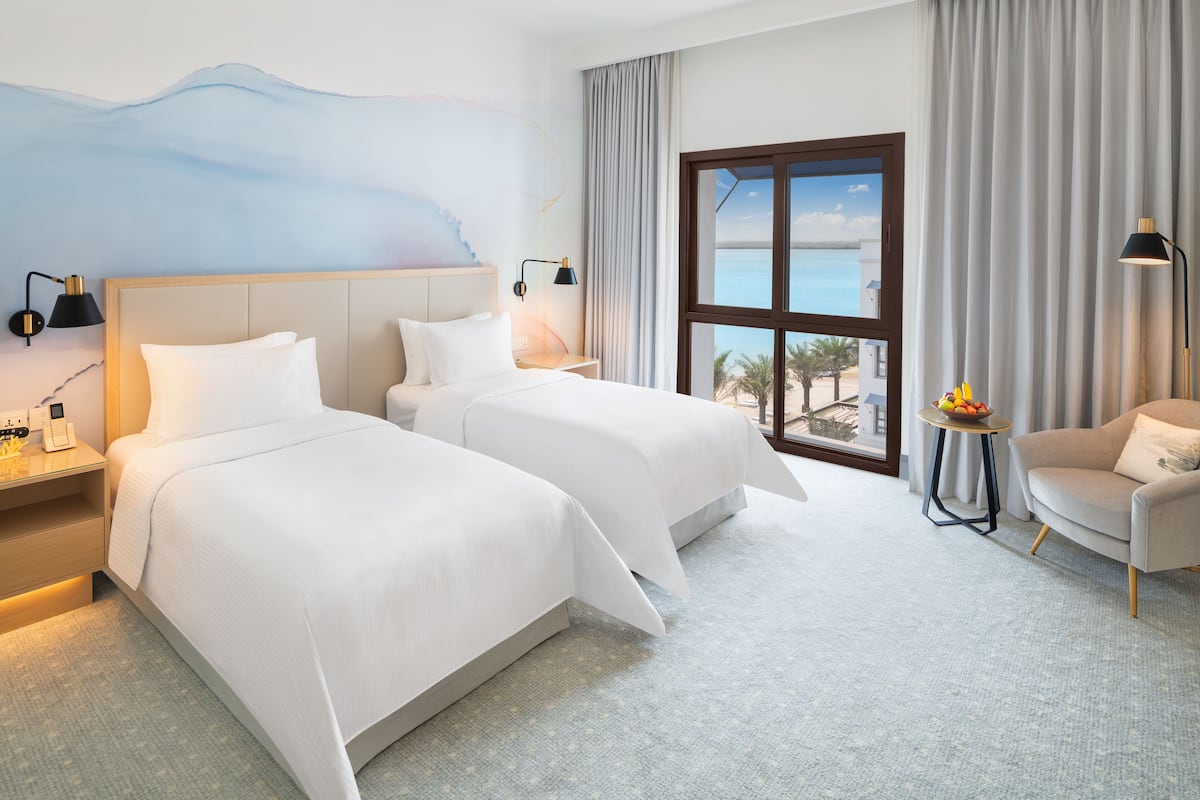 Ein Hotelzimmer mit zwei Betten, einem Sofa, einem kleinen Tisch mit Obst und einem großen Fenster mit Meerblick.