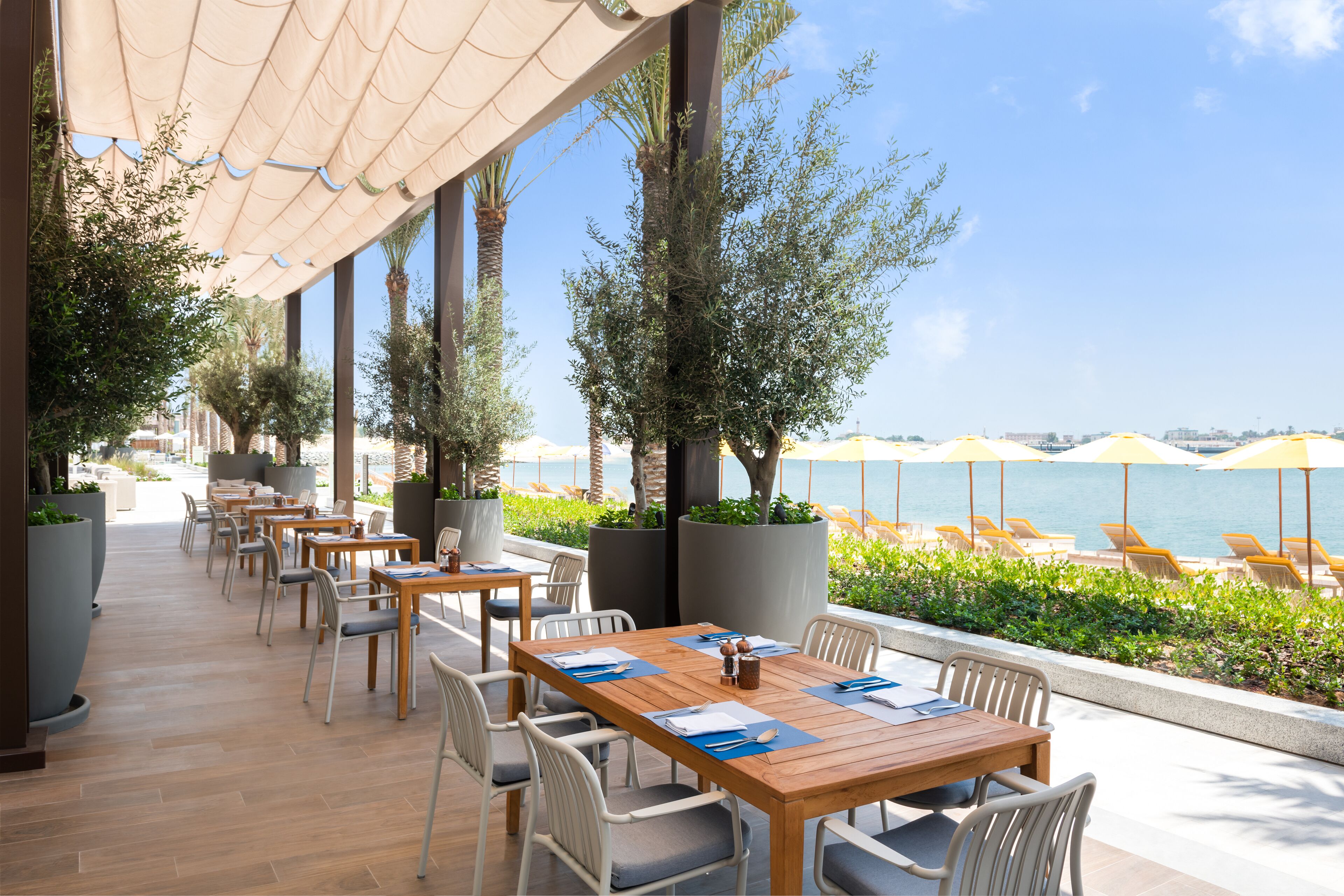 A covered outdoor dining area with tables and chairs, overlooking a beach with umbrellas and lounge chairs.