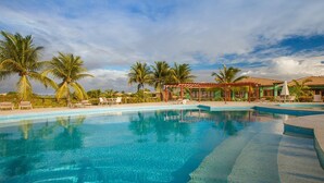 Una piscina cristallina blu con lettini e palme, circondata da un edificio in stile resort con tetto in tegole.